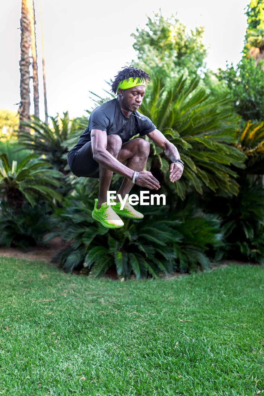 Full body of young african american male athlete jumping high above green lawn while working out in tropical park