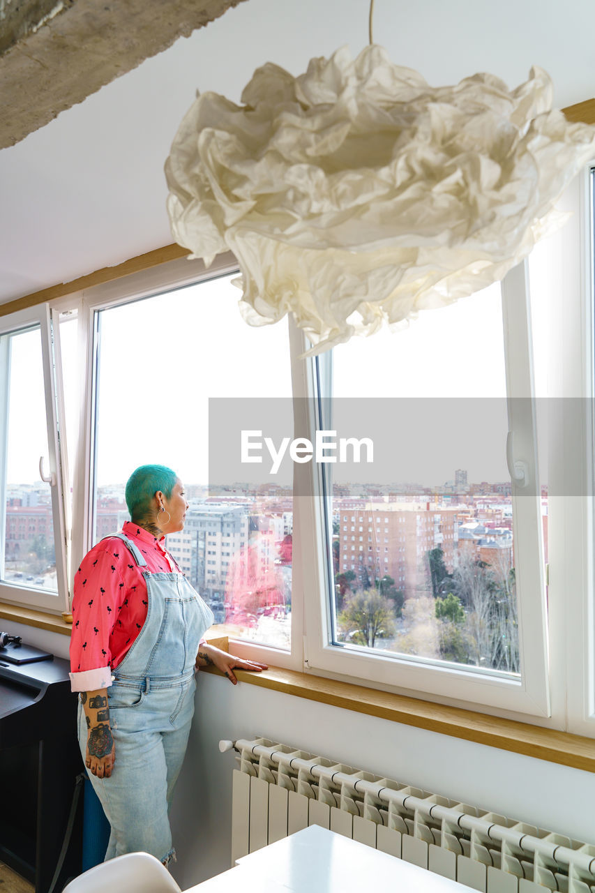 Woman with short dyed green hair looking out window overlooking city with buildings while standing in light room at home