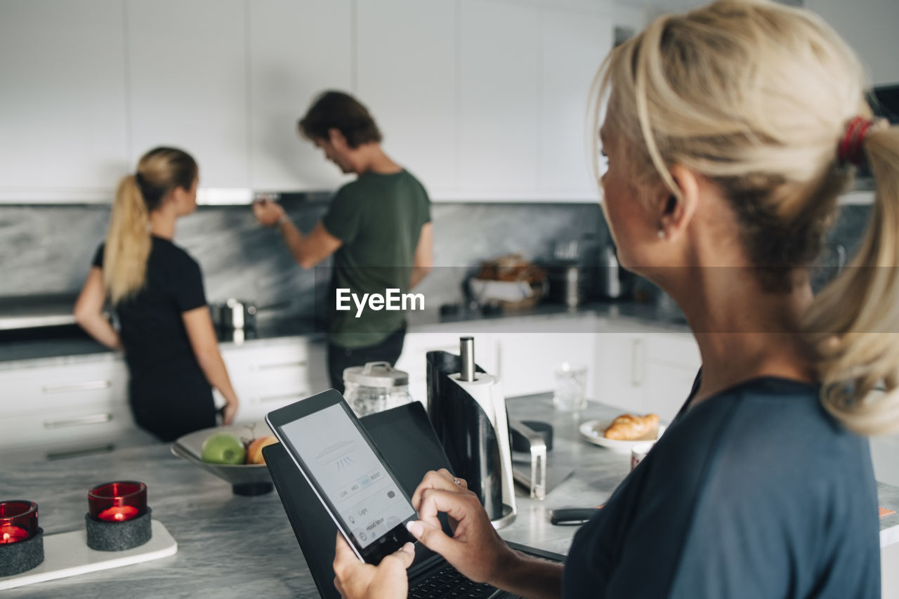 Mature woman with technologies looking at man and daughter standing in kitchen