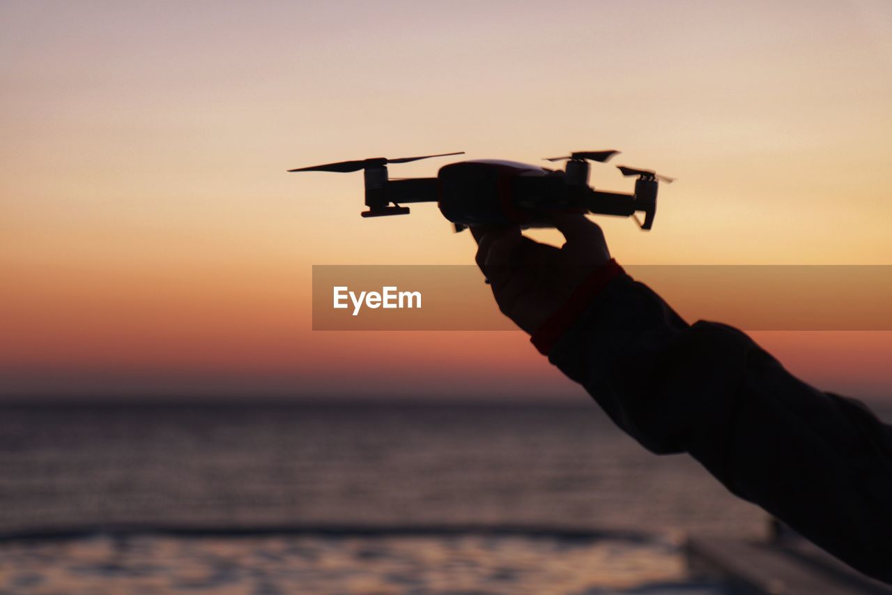 Cropped hand of woman holding drone against sea during sunset