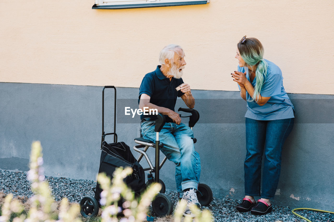 Smiling female nurse clapping while looking at senior man sitting on wheelchair against wall at back yard