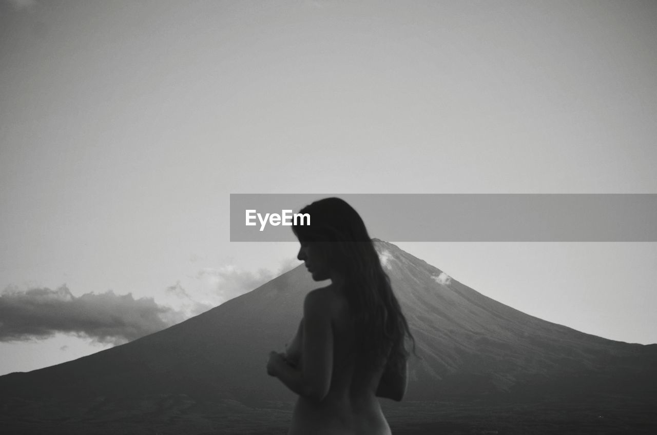Rear view of shirtless woman standing against mountain and sky during dusk