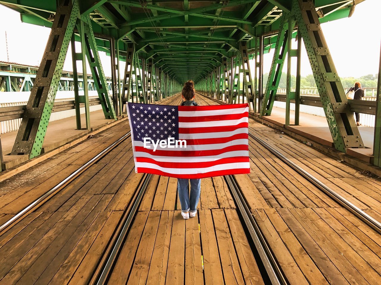 Rear view of woman standing while holding american flag at railway bridge