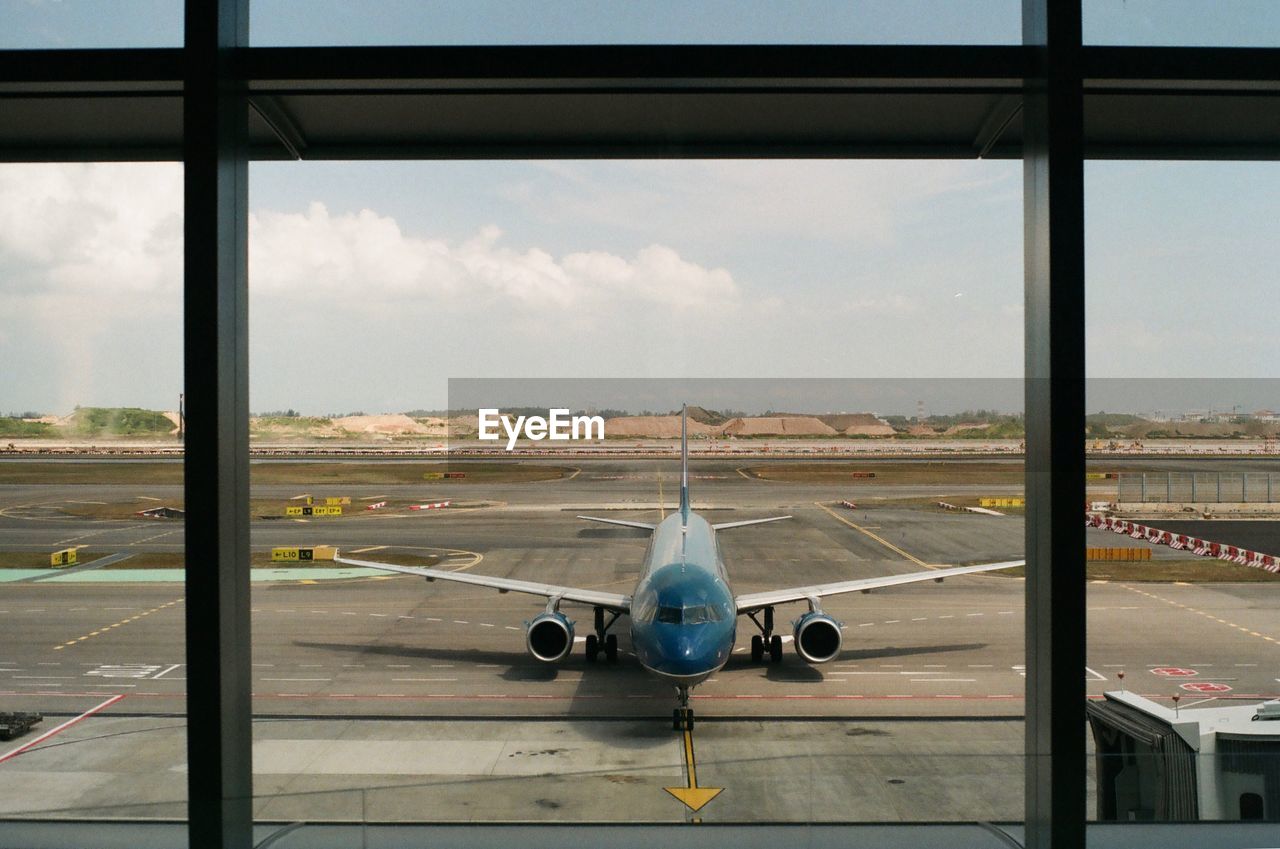 AIRPLANE ON RUNWAY AGAINST SKY SEEN THROUGH GLASS WINDOW