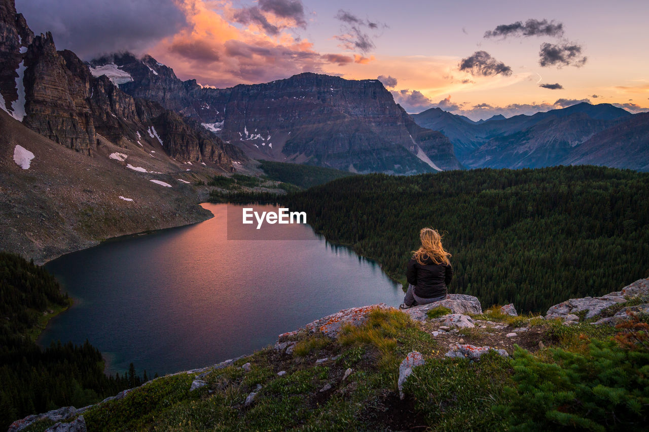 Rear view of woman sitting on cliff by rocky mountains against sky during winter