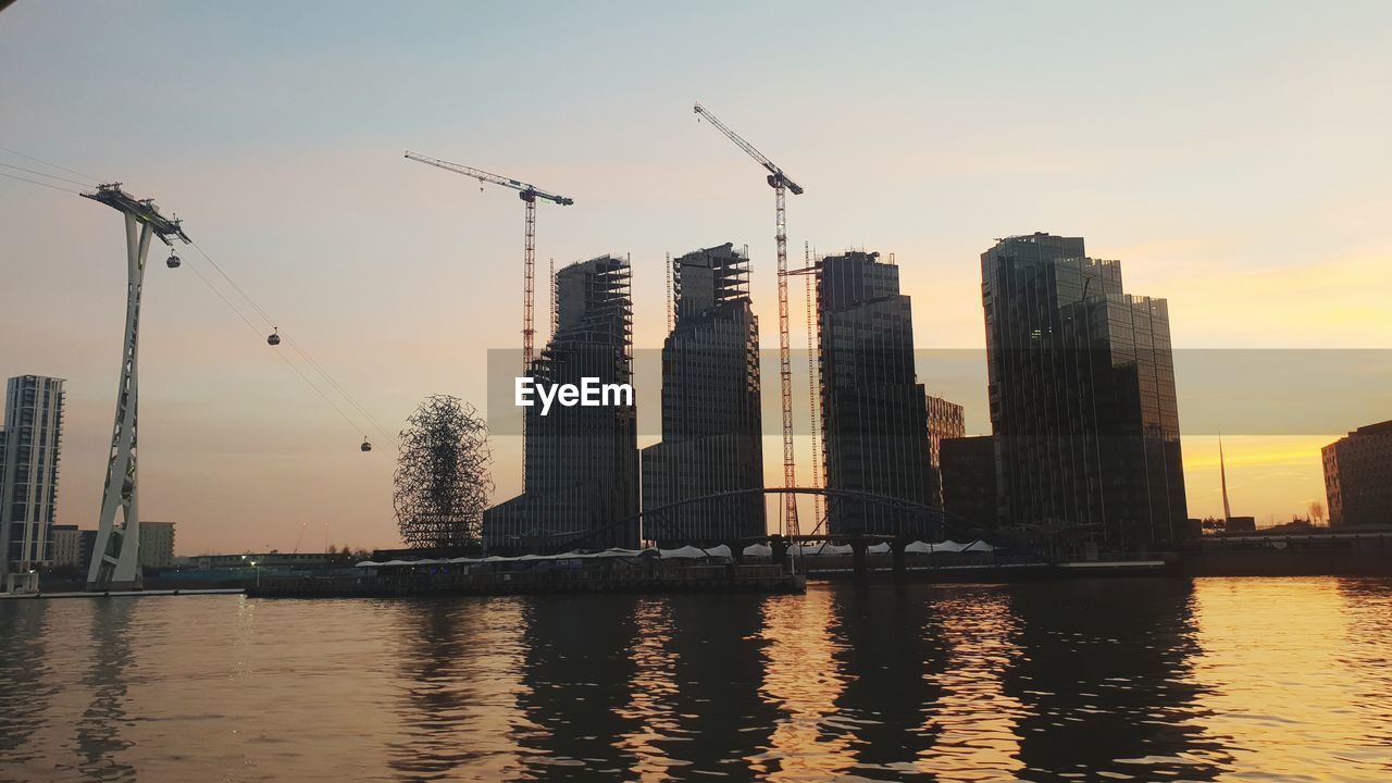 MODERN BUILDINGS BY RIVER AGAINST SKY AT SUNSET