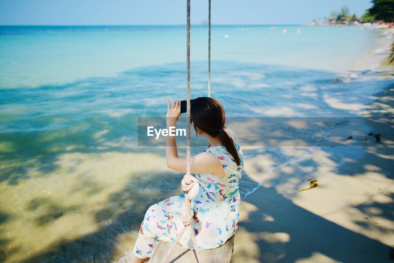 High angle view of woman photographing sea while sitting on swing at beach