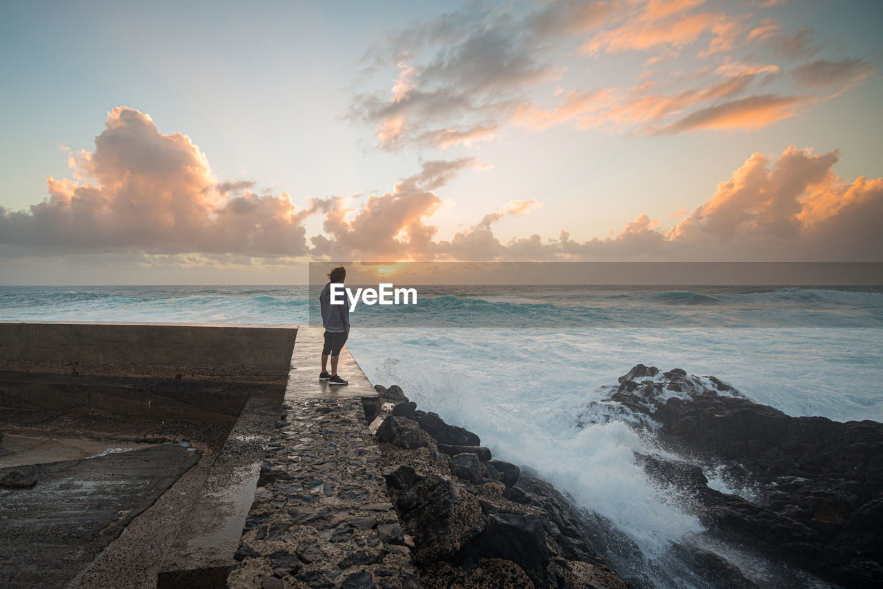 Side view of bearded male with flying hair standing on boulders on coast and observing waving ocean in windy day on fuerteventura island