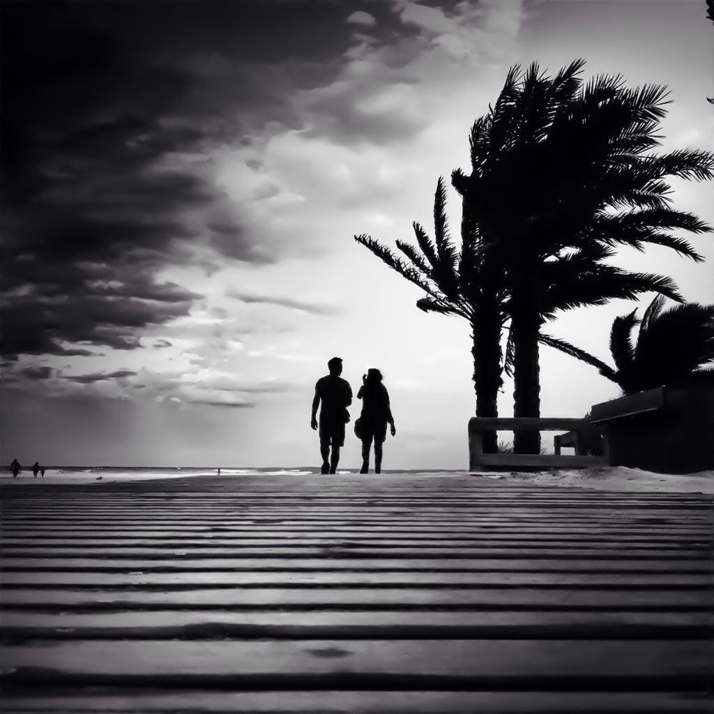 Full length rear view of silhouette couple waking on walkway by beach