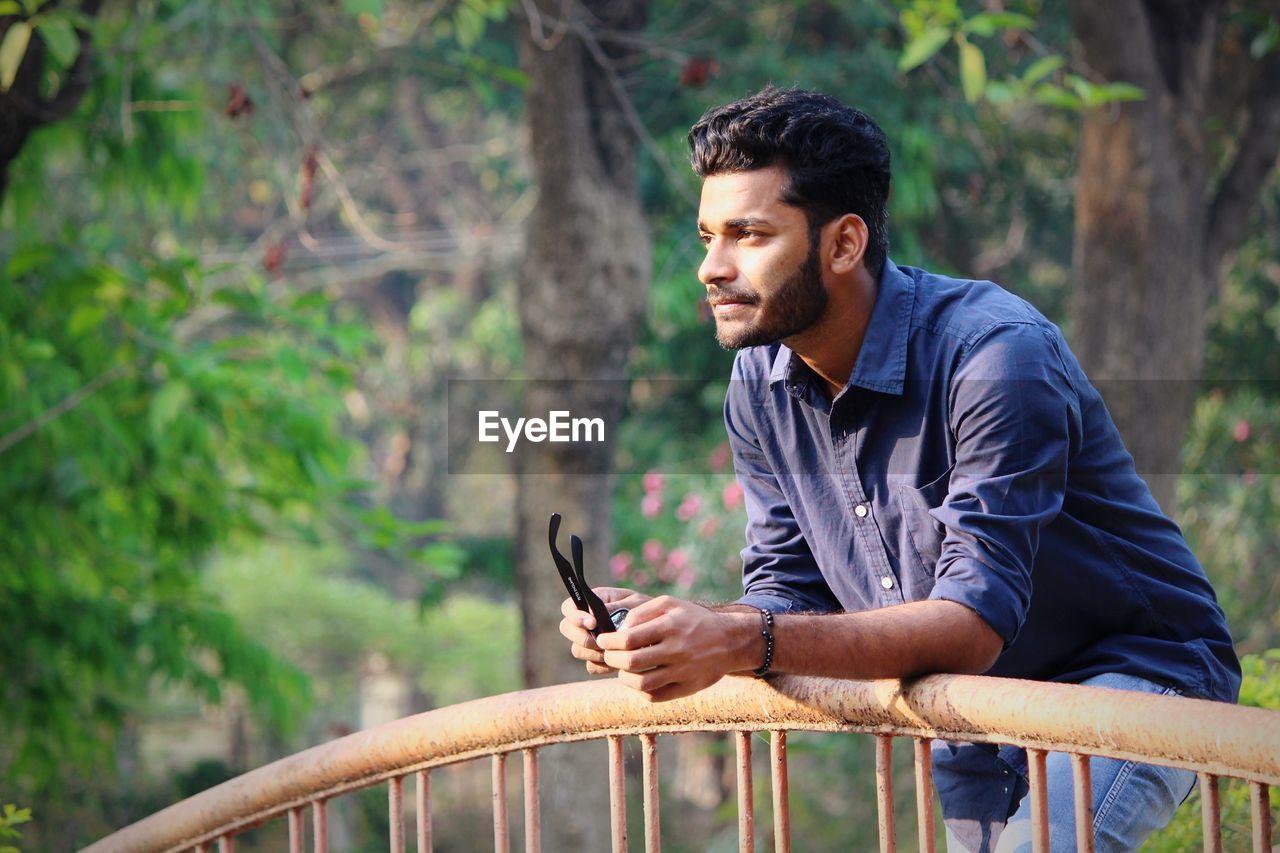 Man looking away while standing by railing against trees