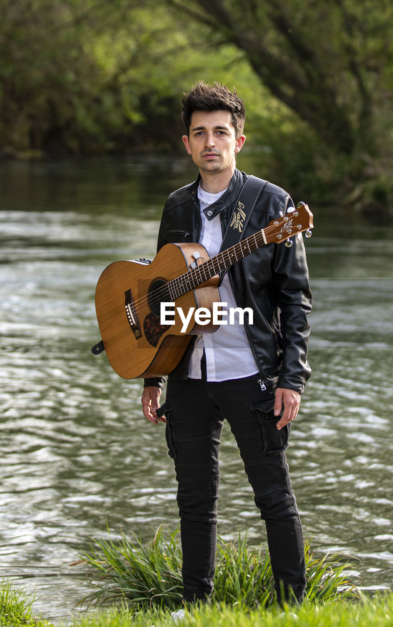 string instrument, musical instrument, music, guitar, one person, musician, young adult, adult, arts culture and entertainment, musical equipment, water, men, front view, full length, nature, standing, portrait, lake, leisure activity, performance, holding, acoustic guitar, day, plucking an instrument, plant, person, casual clothing, outdoors, looking at camera, clothing, skill