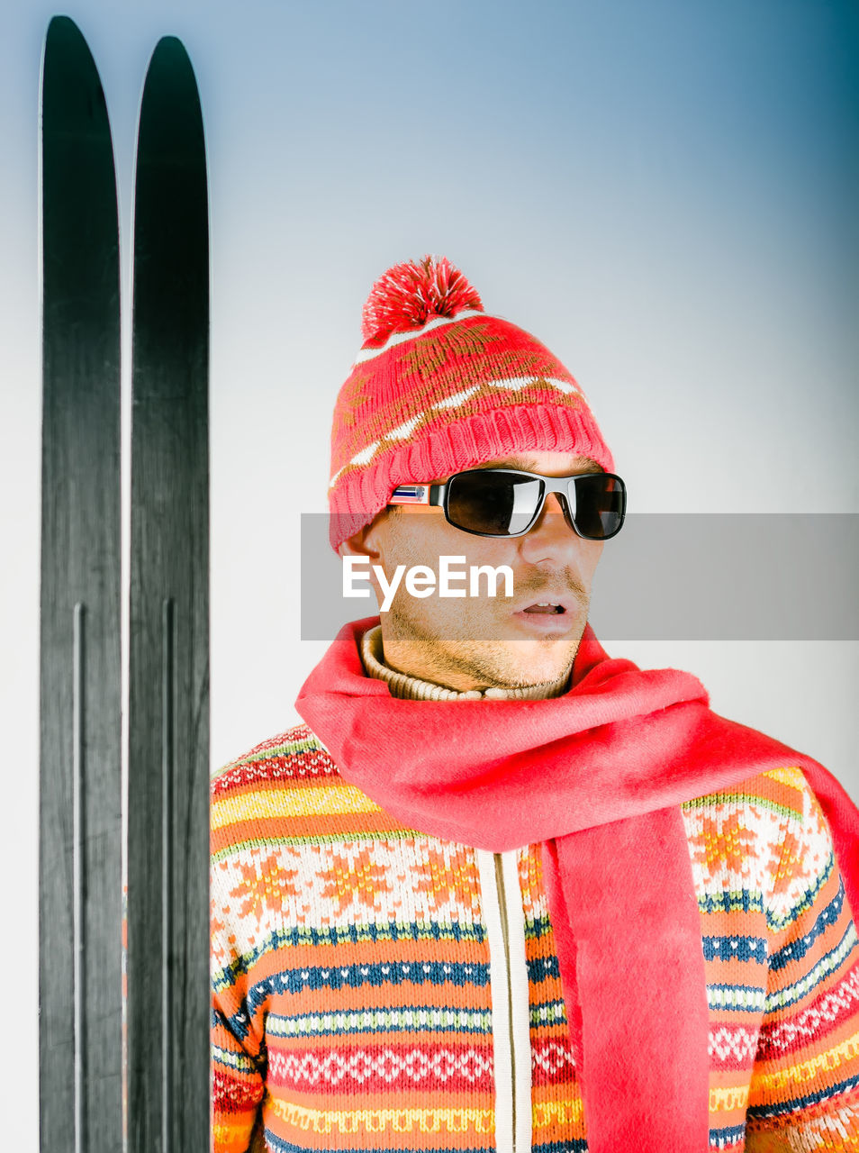 Man in a knitted sweater, woolen cap, scarf and sunglasses stands with skis against blue background.