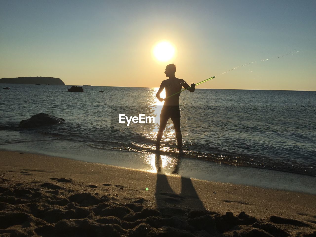 Silhouette man playing with squirt gun at beach against sky during sunset