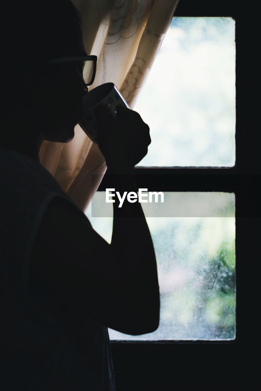 Side view of woman having coffee while standing by window