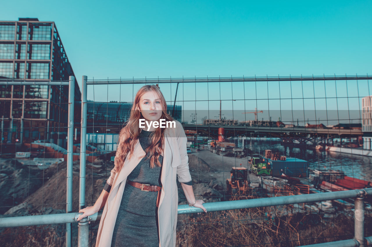 PORTRAIT OF YOUNG WOMAN STANDING ON RAILING AGAINST BRIDGE