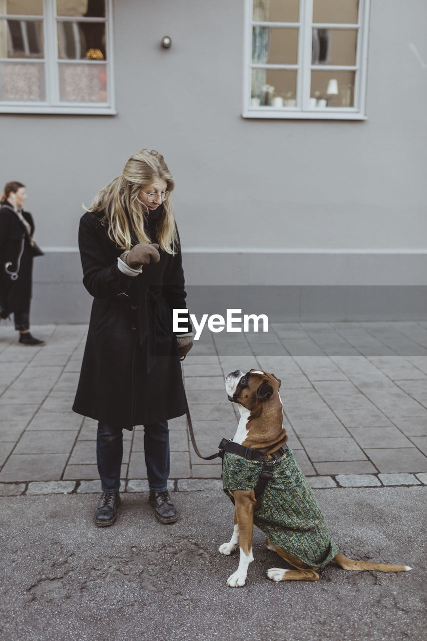 Full length of young woman standing with boxer dog against building in city