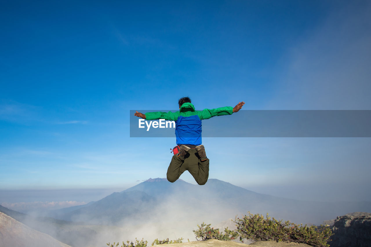 Rear view of man jumping on mountain against blue sky