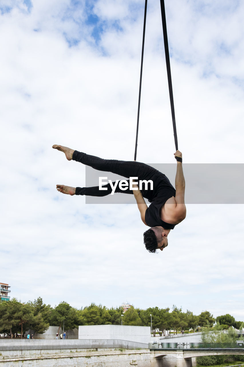 Low angle of fit male gymnast hanging on aerial straps during training in park