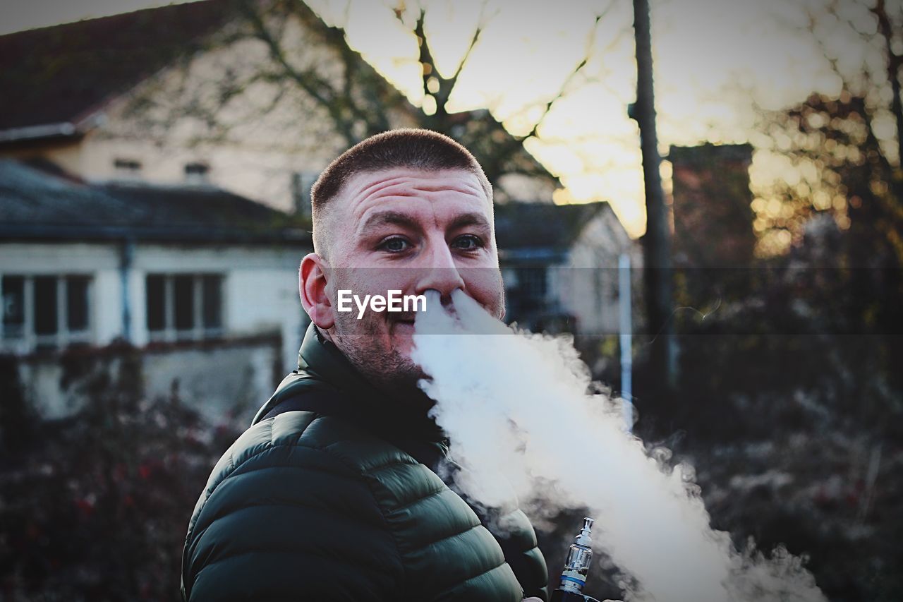 Portrait of young man emitting smoke from nose while standing against house during sunset
