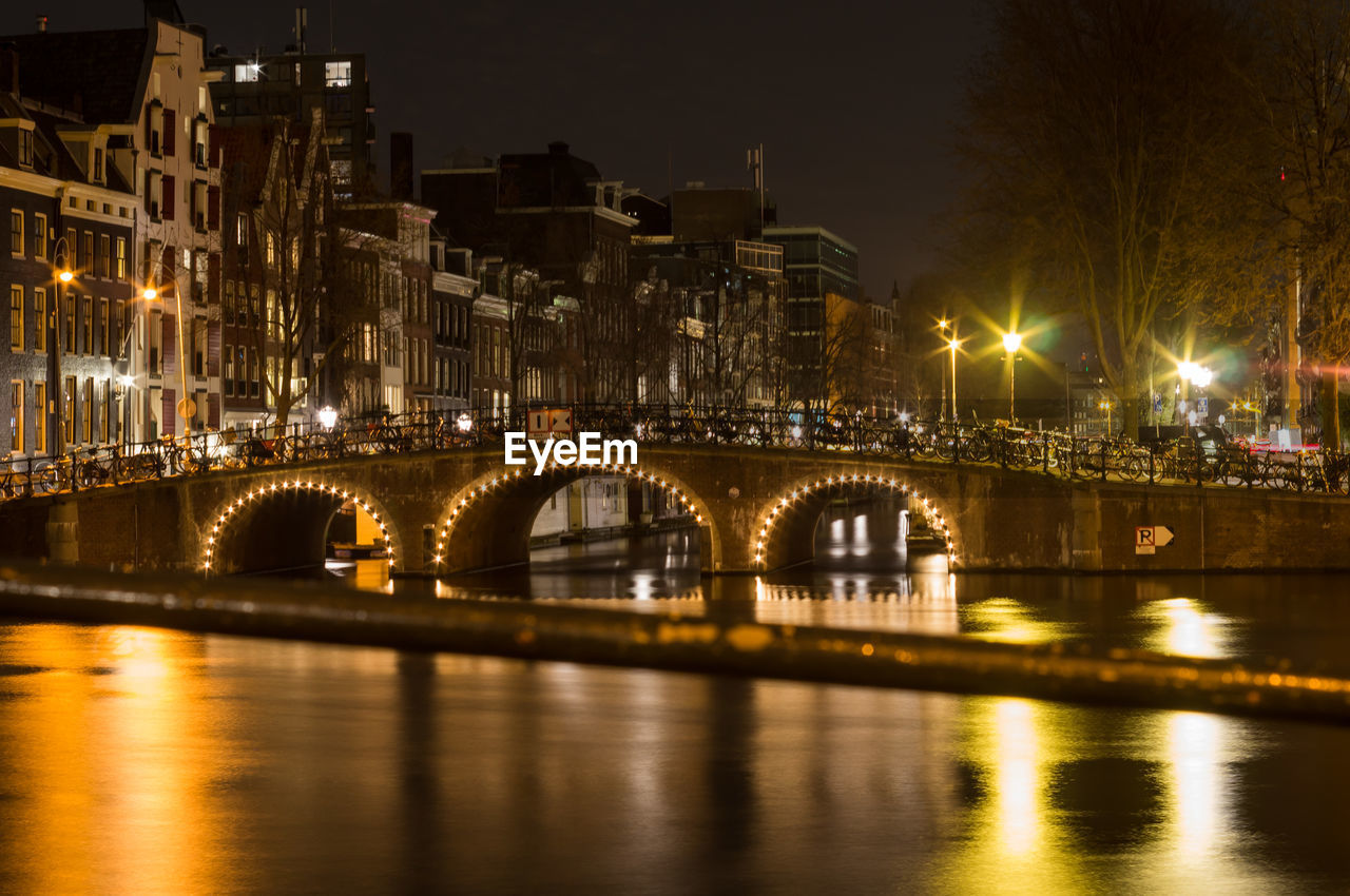 ILLUMINATED BRIDGE OVER RIVER BY BUILDINGS AGAINST SKY