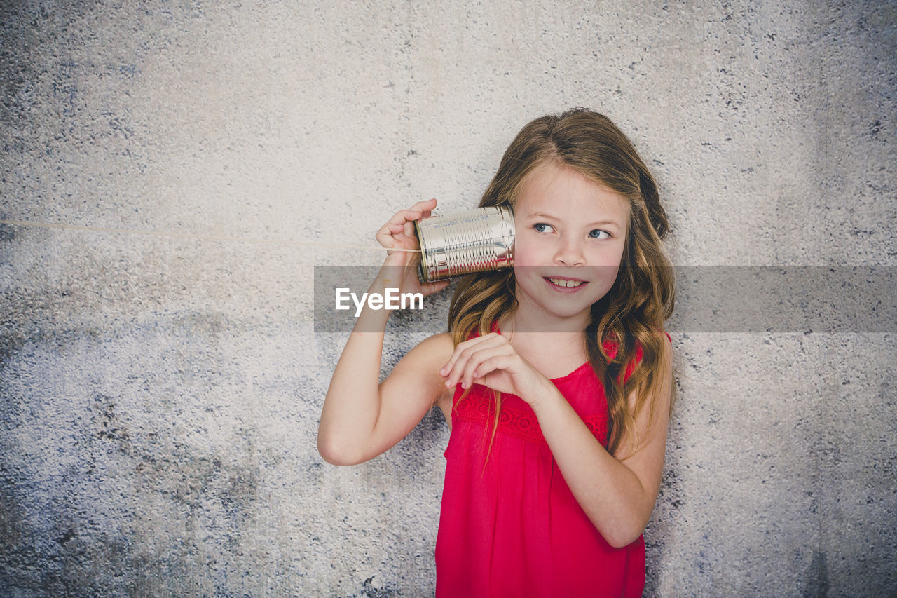 Cute smiling girl playing with tin can phone while standing against wall