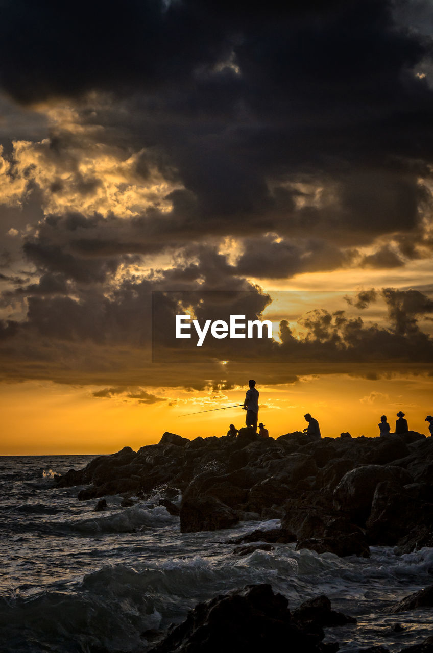 Silhouette people on rock formation in sea against cloudy sky during sunset