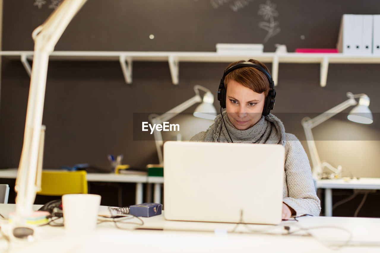 Confident businesswoman listening to headphones while using laptop at creative office