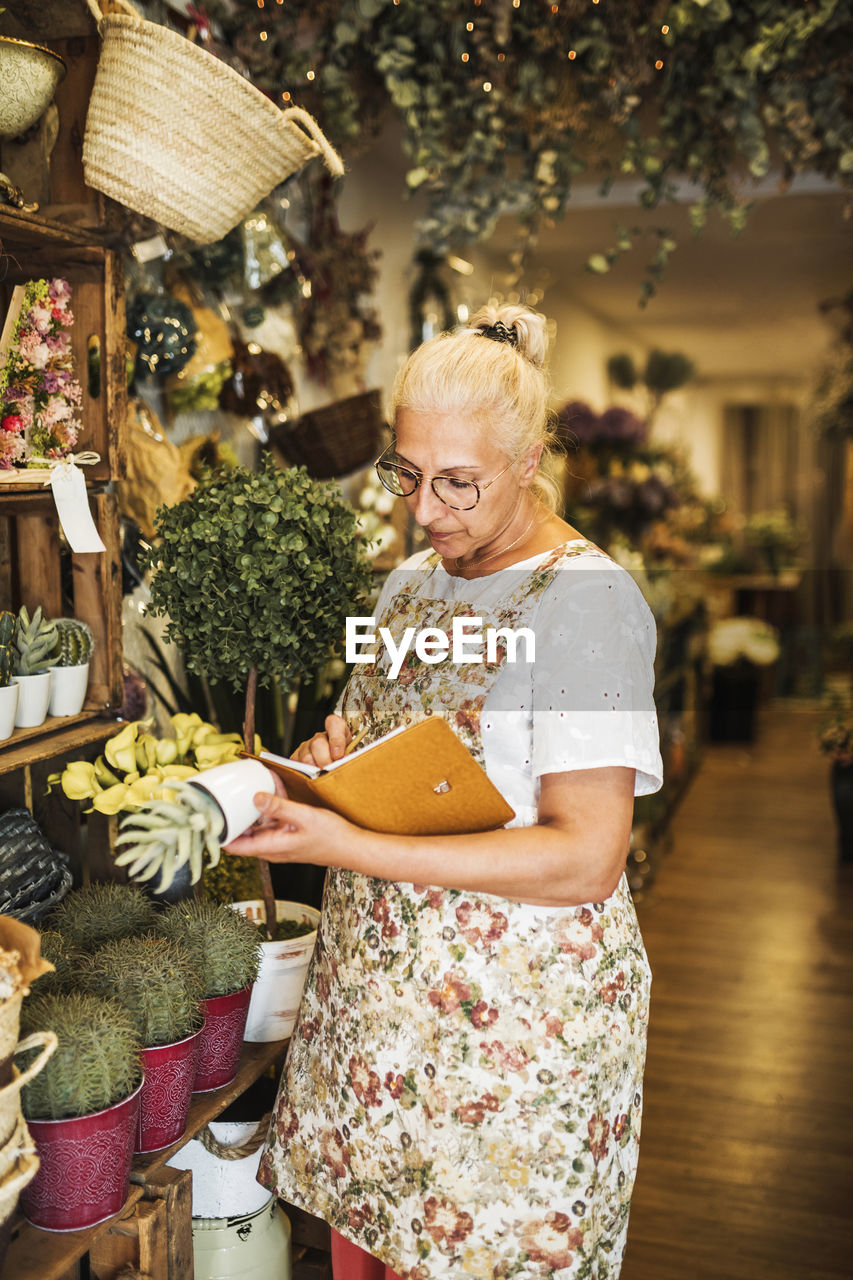 Female florist checking plant while writing in book at store