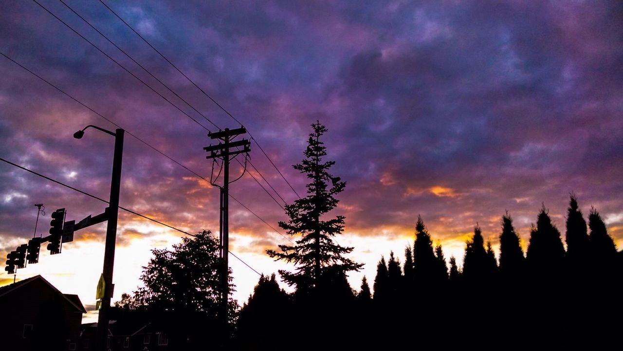 Low angle view of silhouette trees with electricity pylon against cloudy sky during sunset