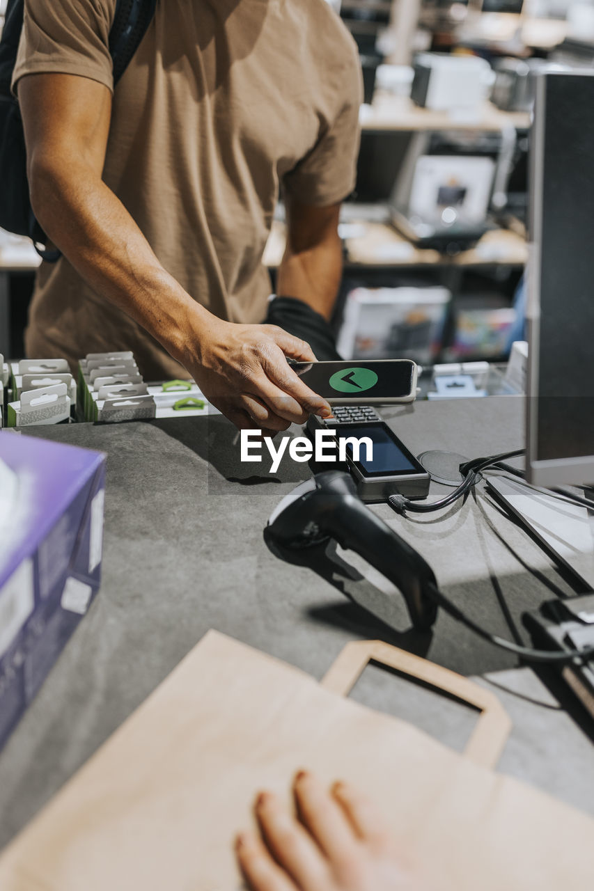 Midsection of male customer scanning credit card reader through smart phone at checkout in electronics store