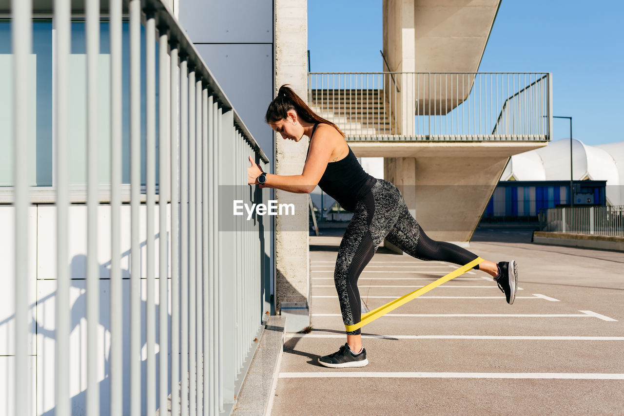 Slim female athlete in sportswear doing exercises with elastic band while standing near metal fence during workout in city resist
