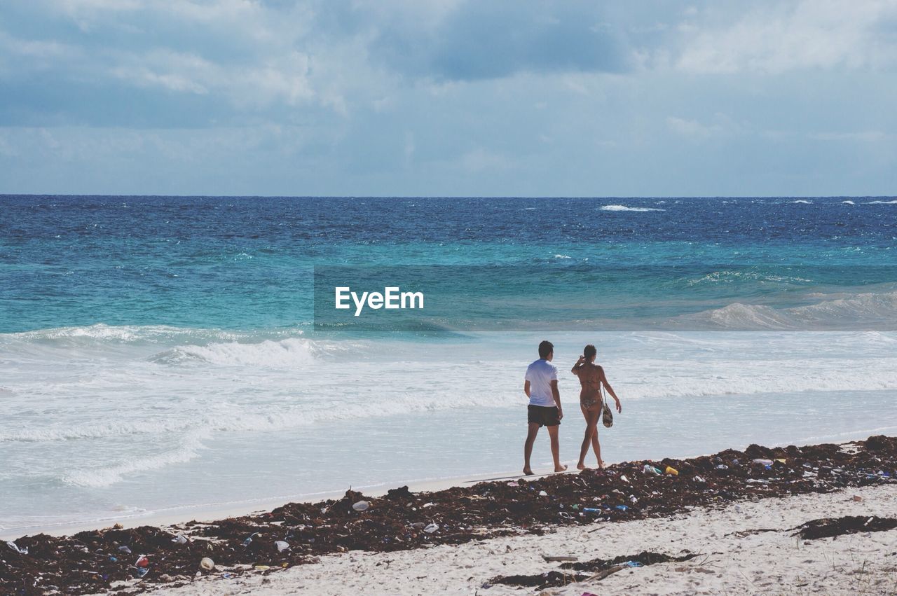 Couple walking on beach against cloudy sky