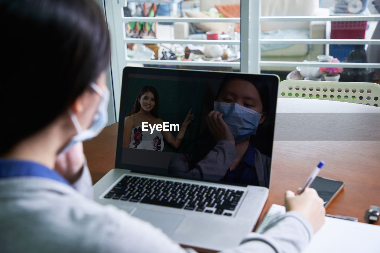 Rear view of girl wearing mask using laptop at home