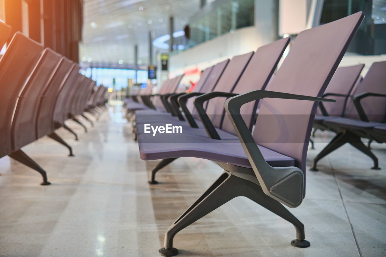 Empty chairs in airport departure hall, blurred airplane in the background.