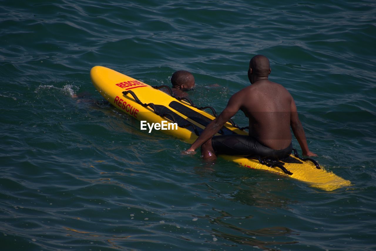 Shirtless man sitting on inflatable raft by friend in sea