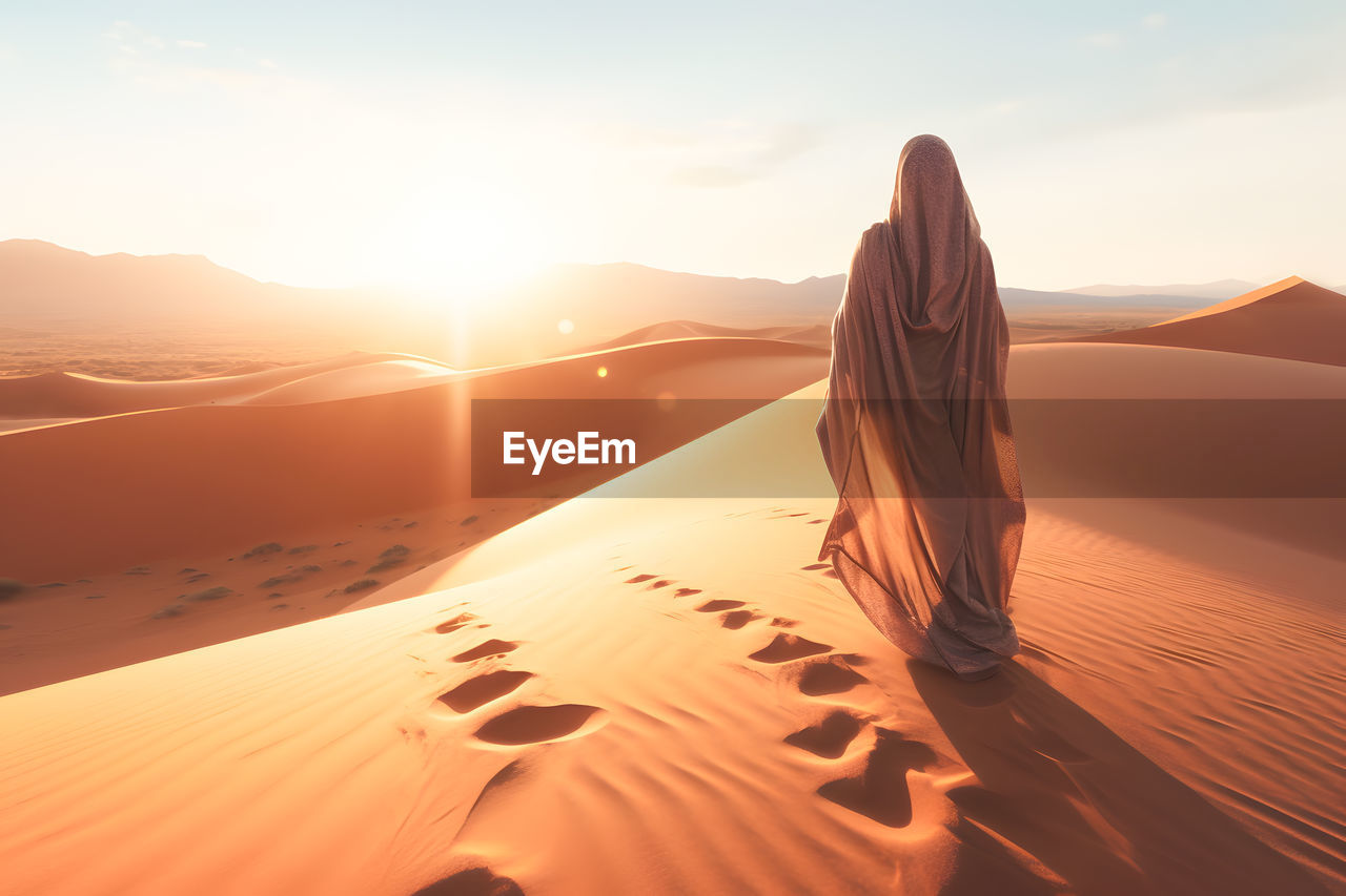 rear view of woman walking on sand at beach against sky during sunset