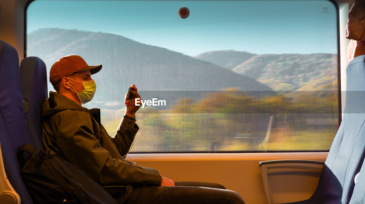 A man in a medical mask with a phone in his hands at a large window on a train.