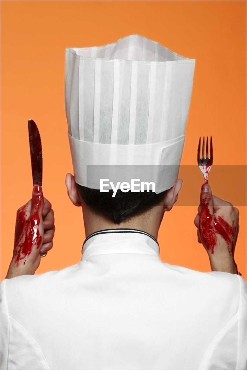 Rear view of chef holding fork and knife with bloody hands against colored background