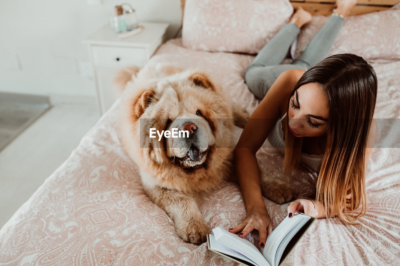 Young woman reading book while lying with dog on bed