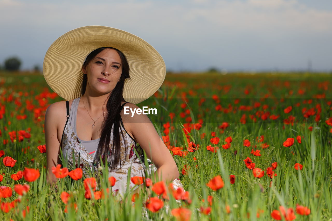 Portrait of young woman sitting amidst poppy flowers on field
