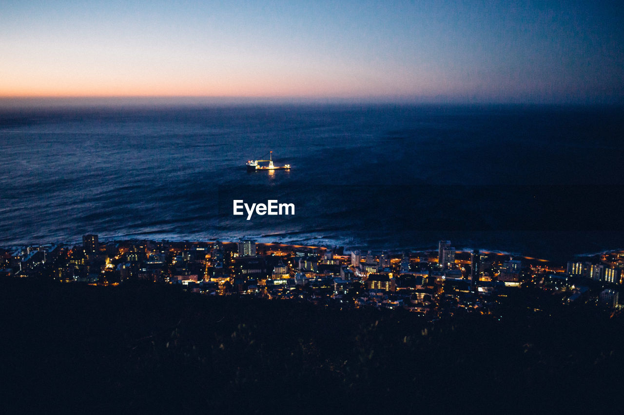 Aerial view of illuminated buildings by sea against sky at sunset