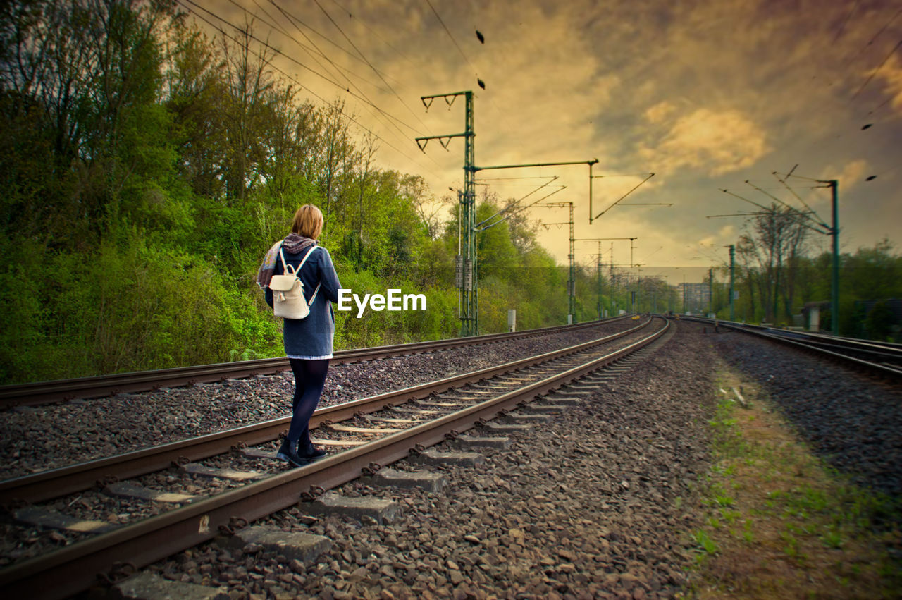 Rear view of backpack woman walking on railroad tracks against cloudy sky