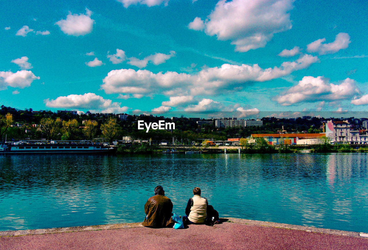 REAR VIEW OF PEOPLE SITTING ON RIVERBANK BY RIVER AGAINST SKY