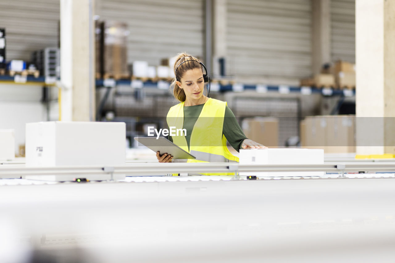 Young worker with tablet pc examining box on conveyor belt in warehouse