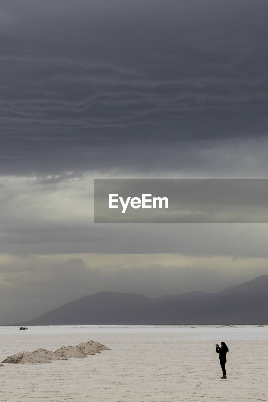 Distant view of woman at salt flat against cloudy sky