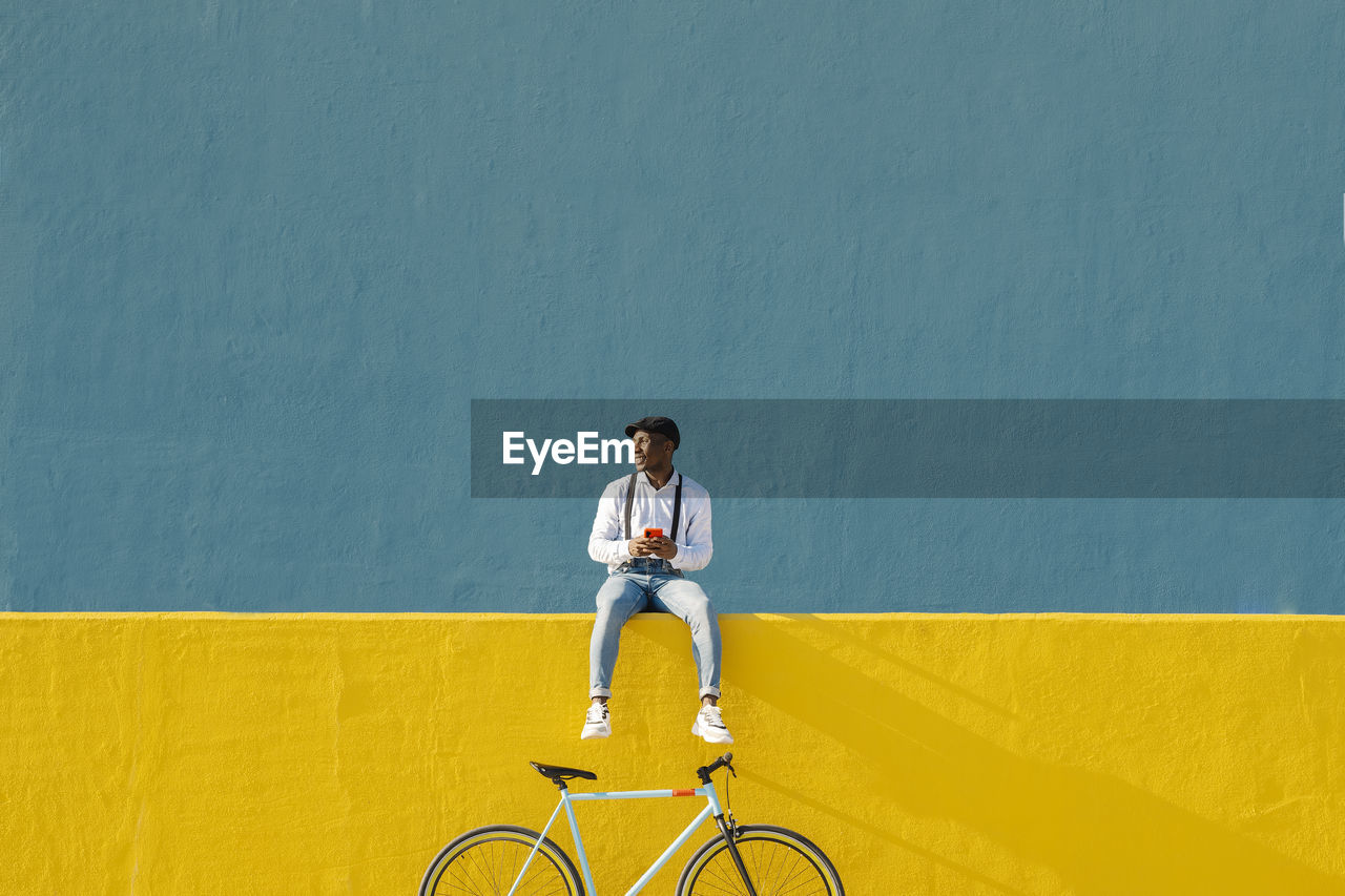 Young man with mobile phone and bicycle sitting on yellow wall