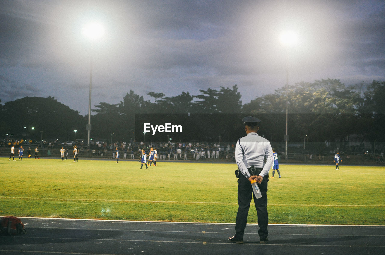 Rear view of security guard looking at players playing soccer on field at night