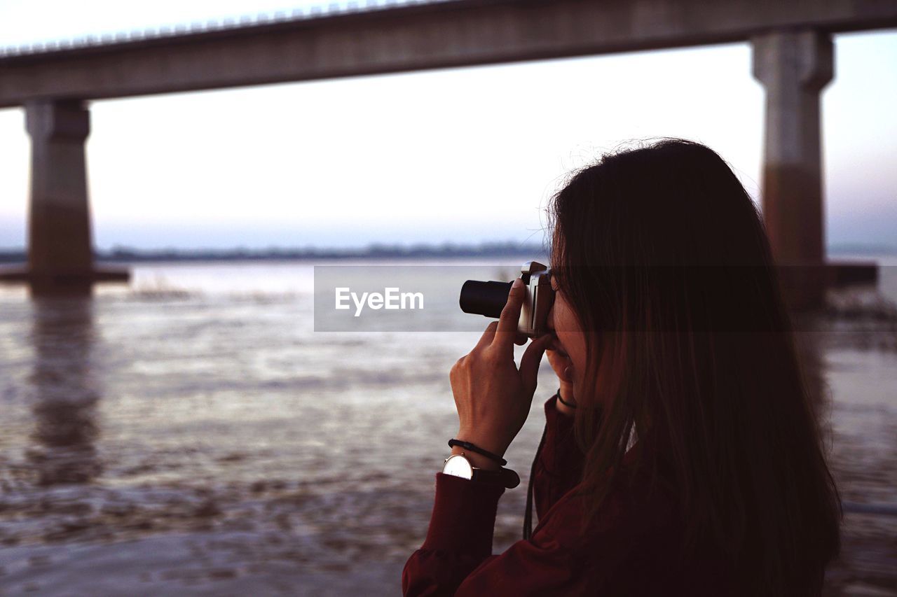 Side view of woman photographing sea through camera during sunset