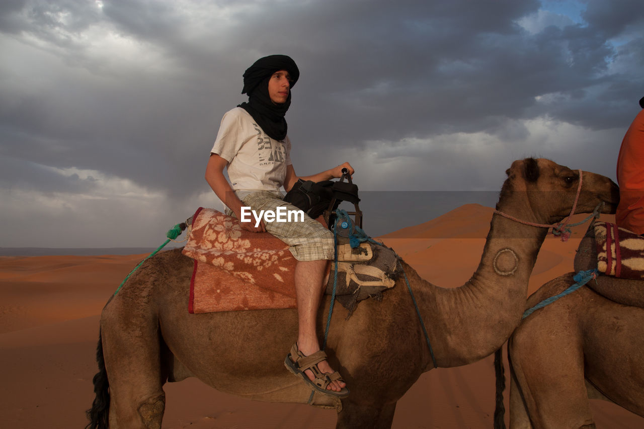 Side view of young man on camel at desert against cloudy sky
