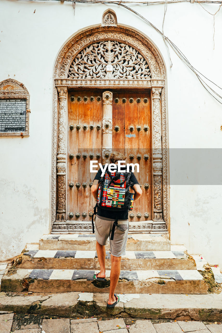 Solo male backpacker with backpack covered in flags stands with ornate door on stone town, zanzibar.